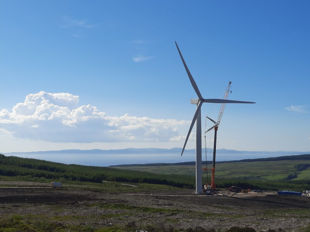 Wind turbine under construction on a hillside with a crane, overlooking green landscape and distant coastline under a clear blue sky